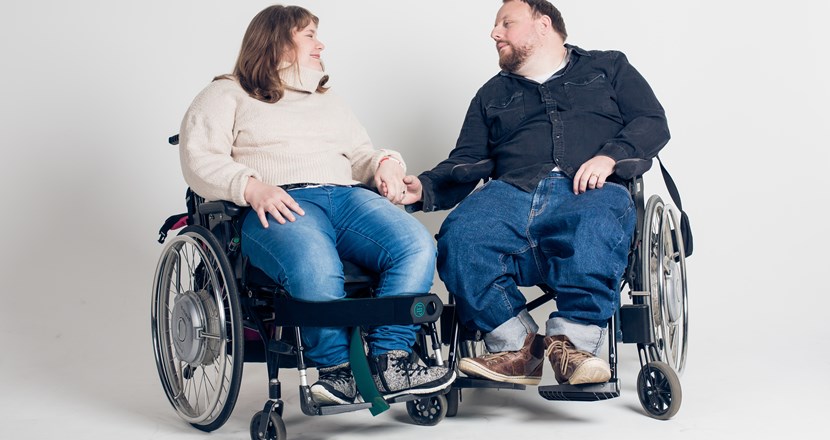 En kvinna och en man i varsin rullstol som håller varandra i handen. Foto.